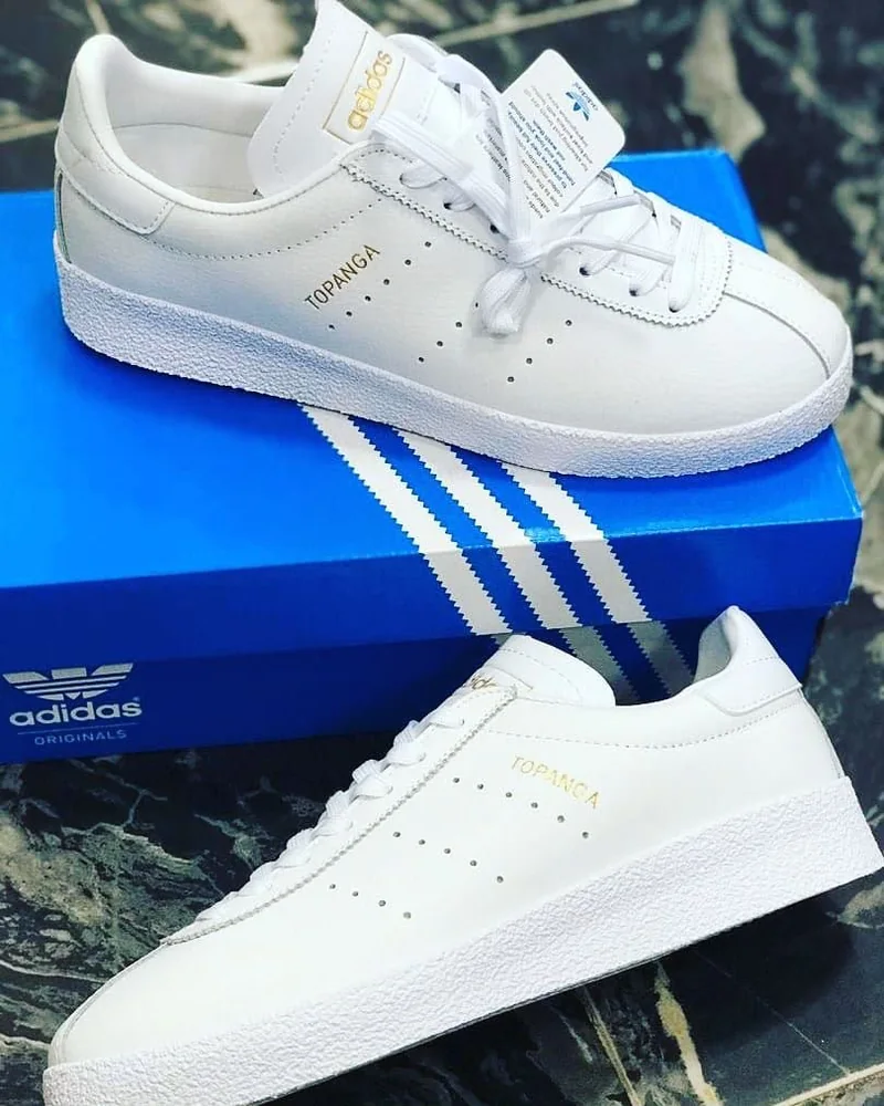 adidas Originals Topanga blanche à détails dorés — Chaussures Oriol Shoes Abomey-Calavi. Basket basse blanche, marquage Topanga doré, perforations latérales, lacets blancs, empeigne semblant en cuir, semelle blanche texturée, présentée sur une boîte adidas bleue