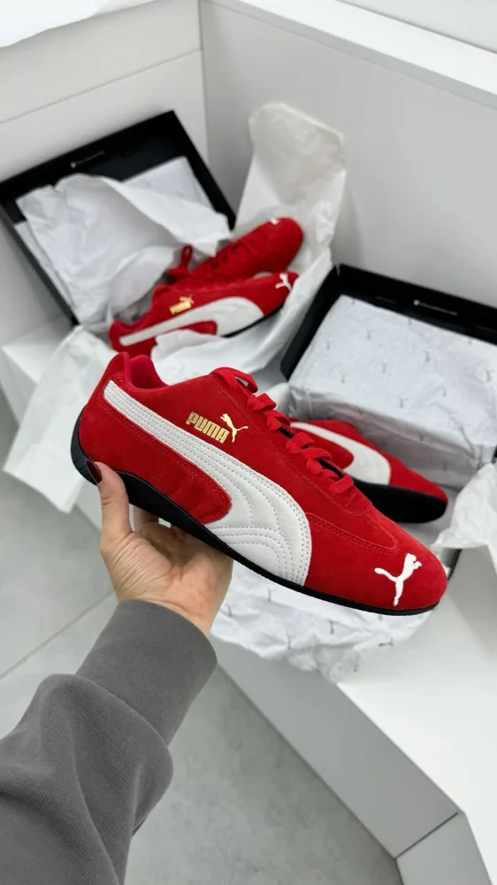 PUMA Speedcat OG For All Time Red / PUMA White — Chaussures Oriol Shoes Abomey-Calavi. Basket basse rouge en matière aspect daim, large bande latérale blanche, logos PUMA blancs et dorés, lacets rouges, semelle noire ; plusieurs chaussures et boîtes visibles en arrière-plan