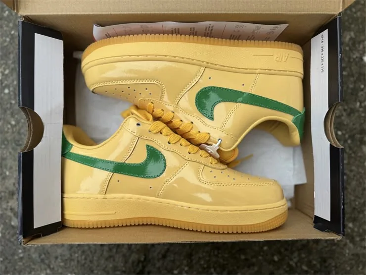 Nike Air Force 1 Low Patent Topaz Gold / Pine Green — Chaussures Oriol Shoes Abomey-Calavi. Paire de baskets basses en cuir ou matière synthétique jaune pâle, grands Swoosh verts, lacets jaunes, semelle ton sur ton et perforations visibles sur l’avant ; présentée dans une boîte à chaussures ouverte