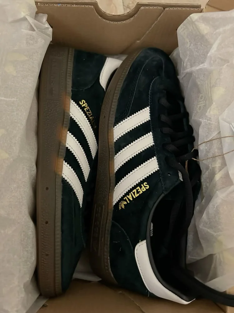 adidas Handball Spezial Core Black / Cloud White / Gum — Chaussures Oriol Shoes Abomey-Calavi. Paire de baskets basses noires, trois bandes blanches, marquage doré « SPEZIAL », empiècements blancs au talon et à la languette, lacets noirs, semelle extérieure gomme marron ; article photographié dans sa boîte