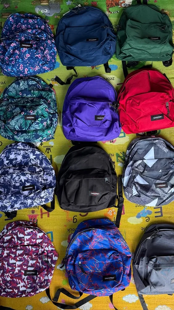 Sacs & bagagerie chez Oriol Shoes Abomey-Calavi — Plusieurs sacs à dos Eastpak visibles, dans des coloris bleu marine, vert, violet, rouge, noir, gris et bleu, avec des versions unies ou à motifs floraux, feuillage et graphiques ; matière textile apparente et poches frontales zippées